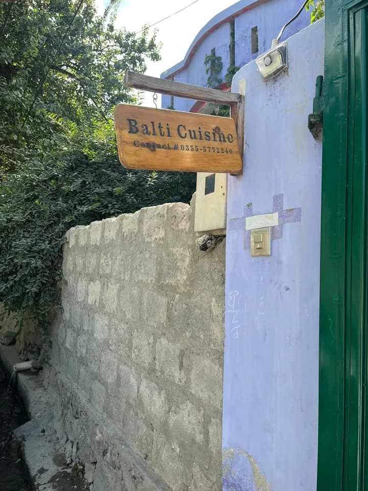 A hand painted wooden sign reading Balti Cuisine, hanging on a lilac wall beside a green gate