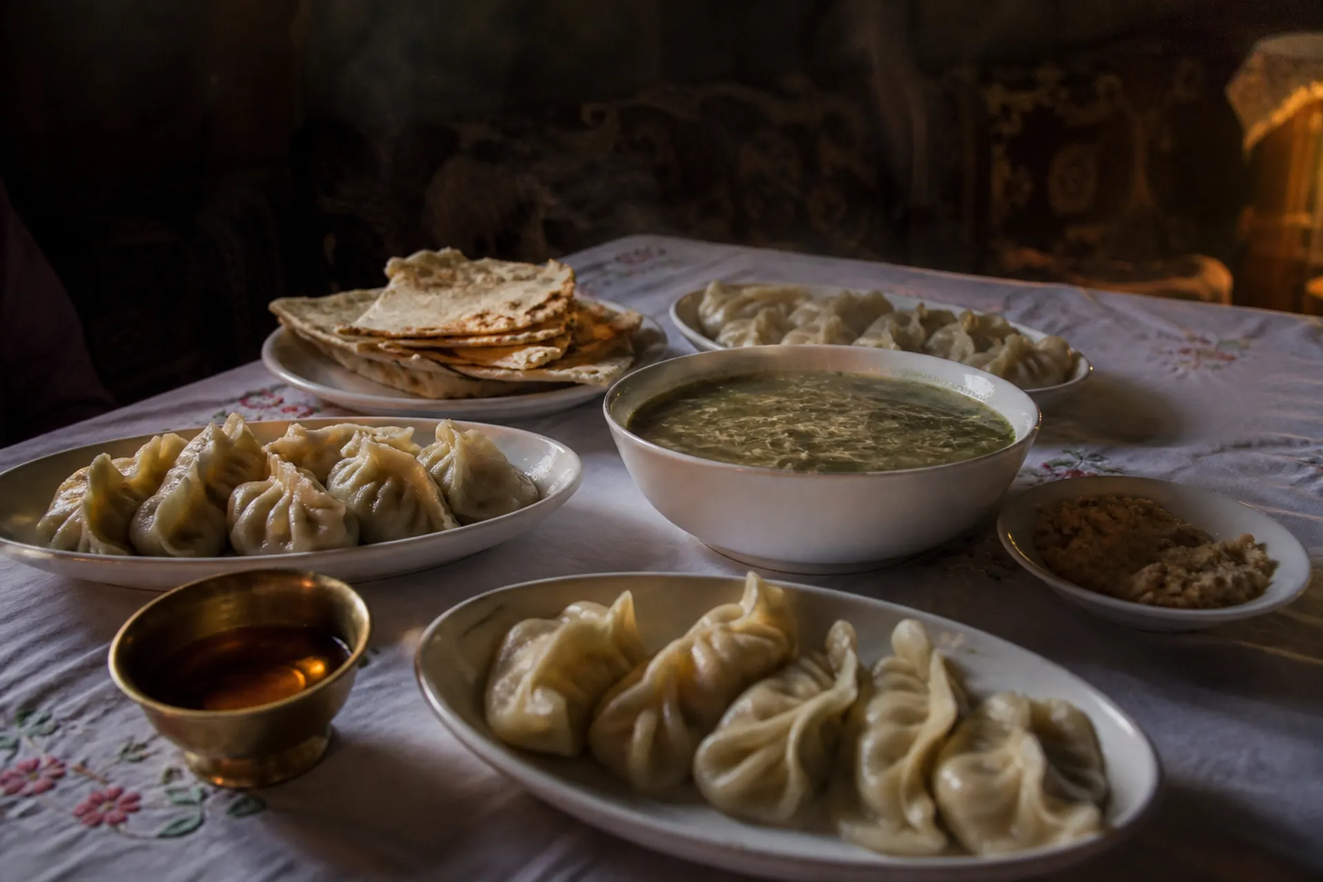 A Balti meal laid on a white cloth: plates of mamtu, a bowl of tras balay, flatbread and a brass dish of apricot oil