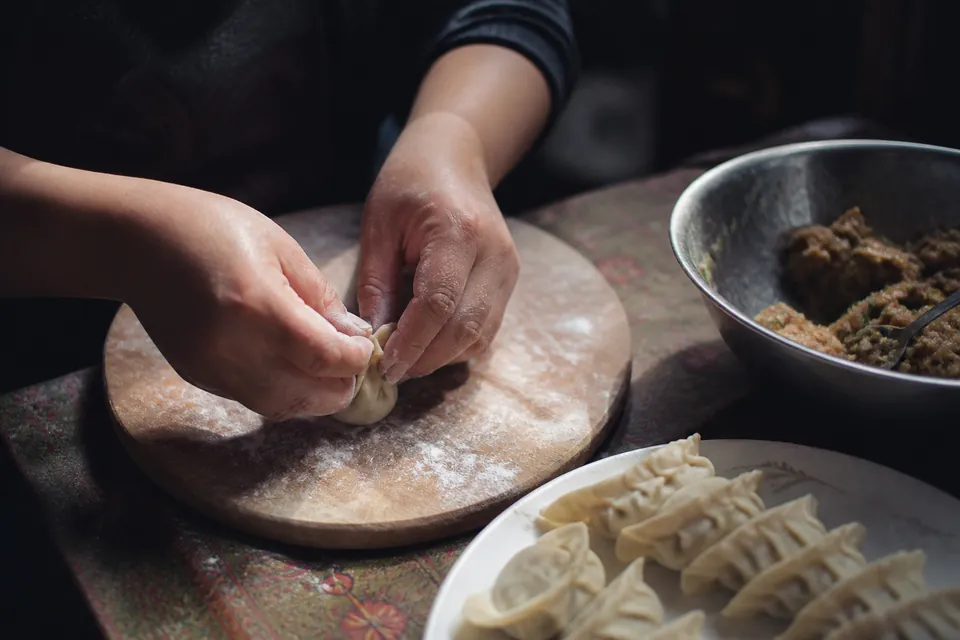 Hands pleating a mamtu over a floured board, a bowl of filling beside them